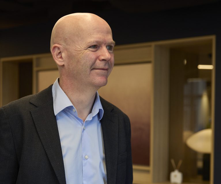 Bald person in a light blue shirt and dark blazer stands indoors, smiling softly, with a modern interior featuring warm lighting and wooden panels behind.