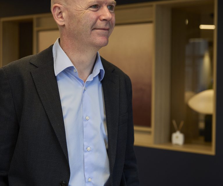 Bald person in a light blue shirt and dark blazer stands indoors, smiling softly, with a modern interior featuring warm lighting and wooden panels behind.