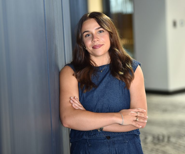 A young woman with shoulder-length brown hair, wearing a sleeveless denim dress, stands with arms crossed, leaning against a grey wall in an indoor setting.
