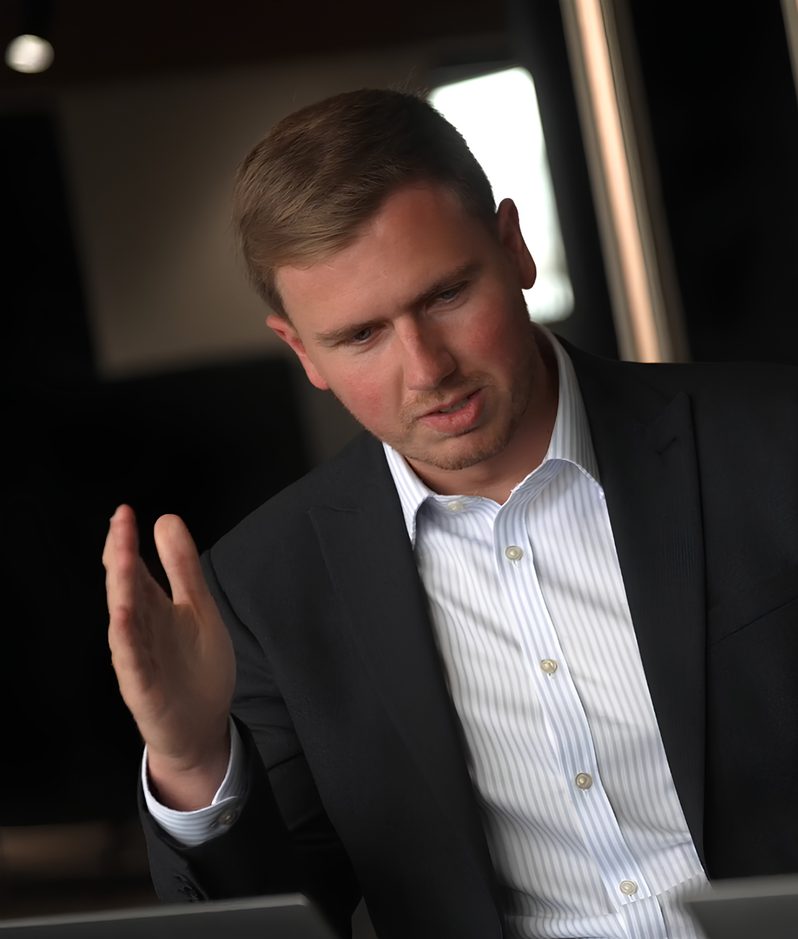 A person in a dark suit and striped shirt sits at a desk, gesturing with an open hand while looking down, in a modern office with a blurred background.