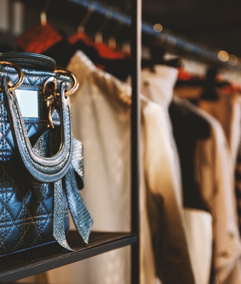 Close-up of a quilted blue handbag with metallic rings on a shelf in a clothing store, with blurred jackets hanging in the background.