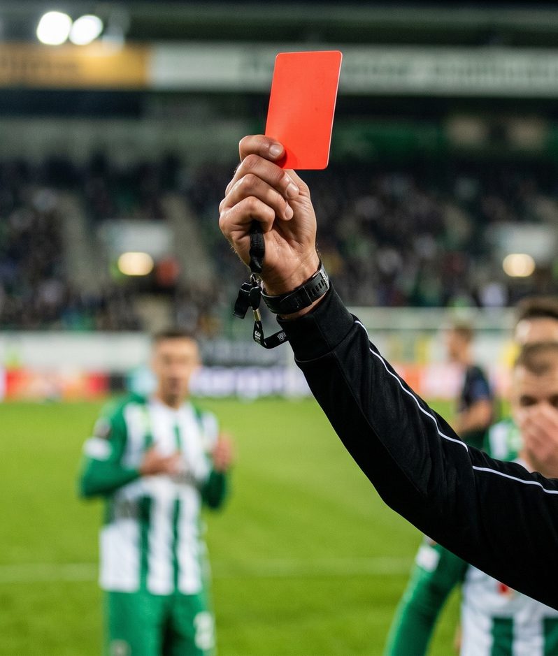 Referee holding up a red card in the foreground during a football match, with players in green and white stripes and a blurred crowd in the stands.