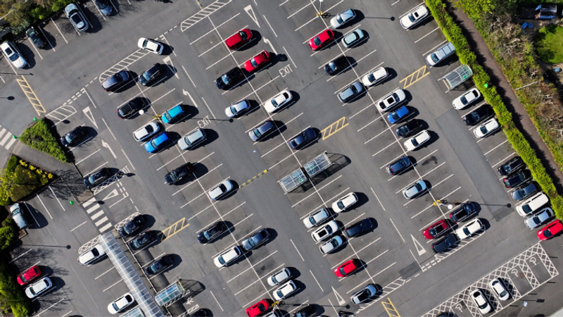 Aerial view of a large parking lot filled with cars in neat rows, white lines marking spaces, zebra crossings, and green hedges along the edges.