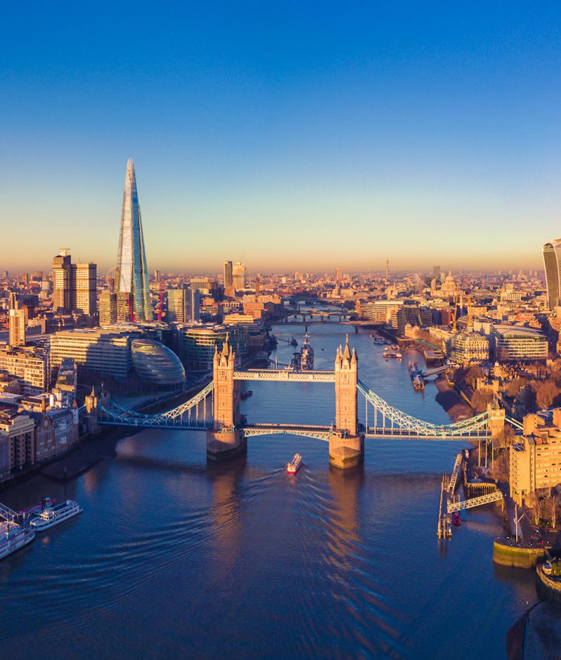 A panoramic aerial view of London at sunset, showcasing the River Thames, Tower Bridge, The Shard, and an array of skyscrapers against a clear blue sky.