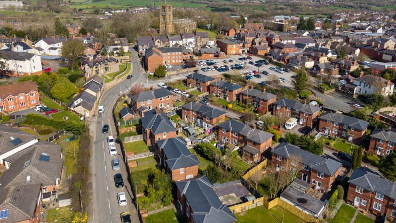 Aerial view of a UK town with dense red-brick houses, winding streets, a prominent church tower, and a central car park surrounded by greenery.