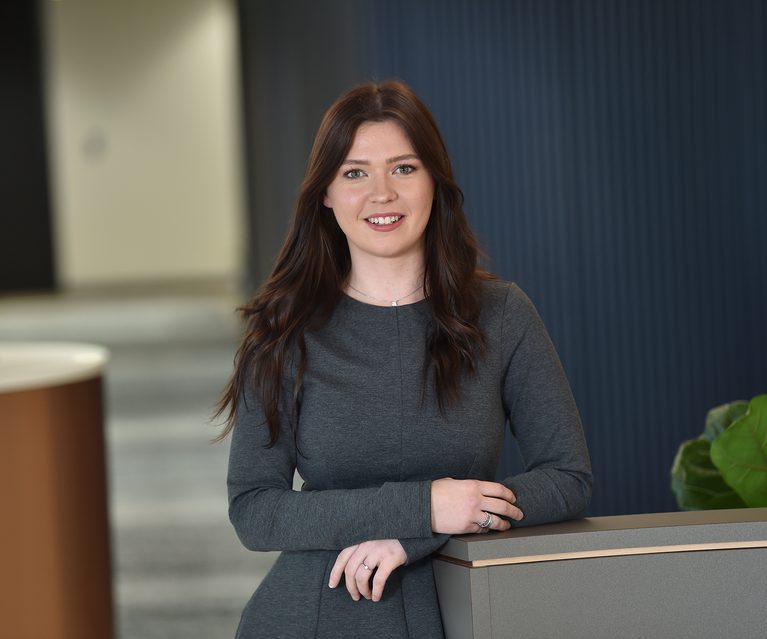 A young woman with long, wavy brown hair and fair skin, wearing a dark grey dress, standing indoors against a blue panel background, smiling gently at the camera.