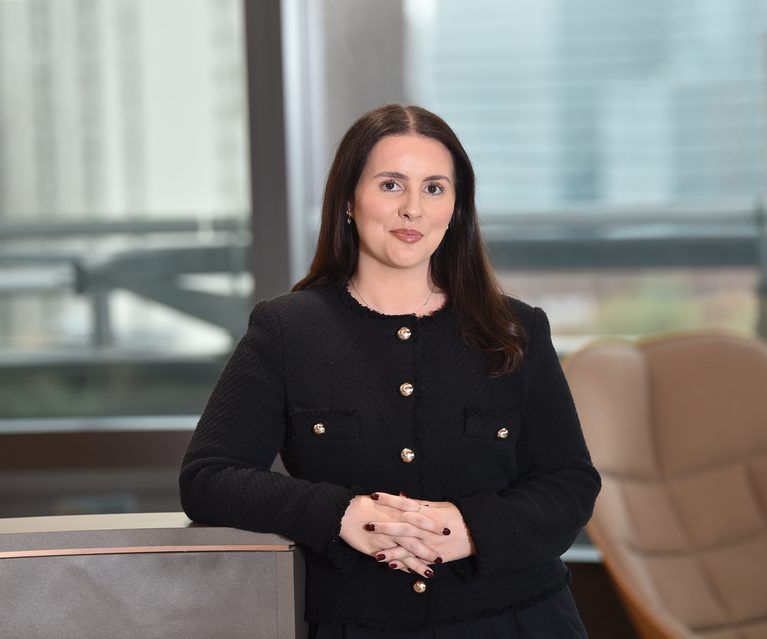 A woman with long brown hair, wearing a black jacket with gold buttons, stands with hands clasped in front of her in a modern office setting.