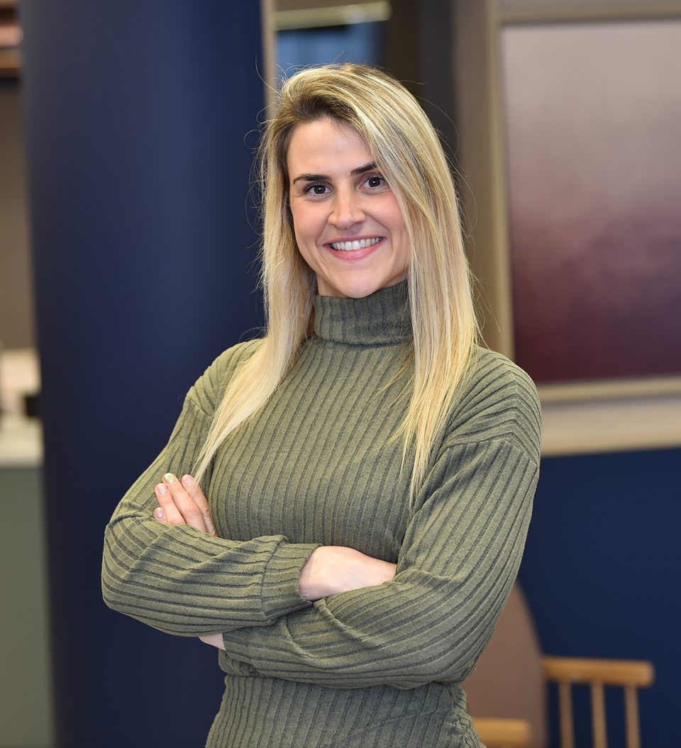 A woman with long blonde hair, wearing a green ribbed turtleneck sweater, smiles with arms crossed in an indoor setting.