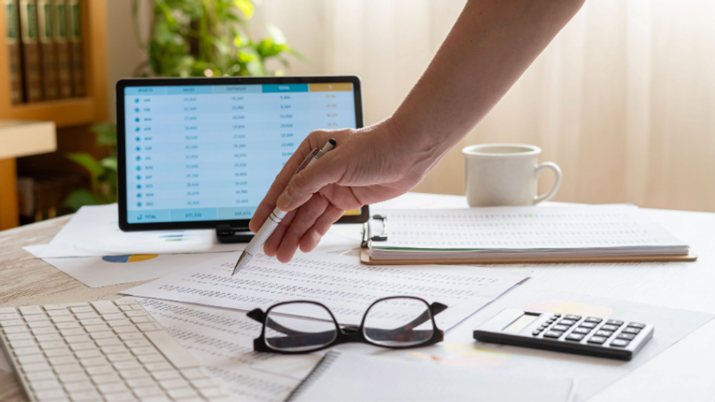 A hand holds a pen over scattered documents on a desk with a laptop showing spreadsheets, glasses, a calculator, clipboard, mug and a plant in the background.