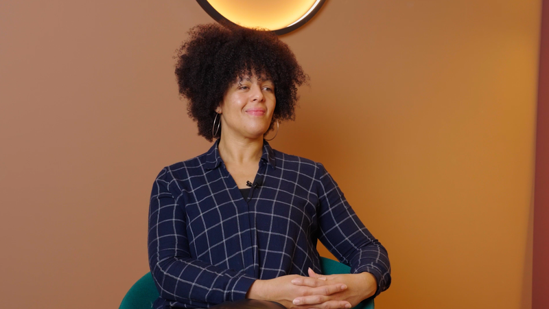 A woman with curly hair wearing a dark checkered shirt sits with her hands clasped, smiling softly against a warm-coloured background.