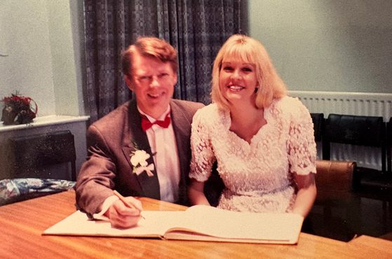Two people sit at a table signing a book; one wears a brown suit with a red bow tie and boutonniere, the other wears a white lace dress, both smiling.