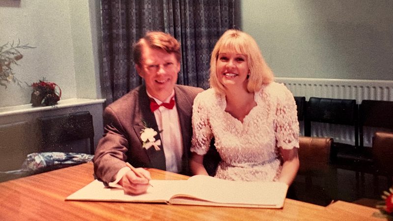 Two people sit at a table signing a book; one wears a brown suit with a red bow tie and boutonniere, the other wears a white lace dress, both smiling.