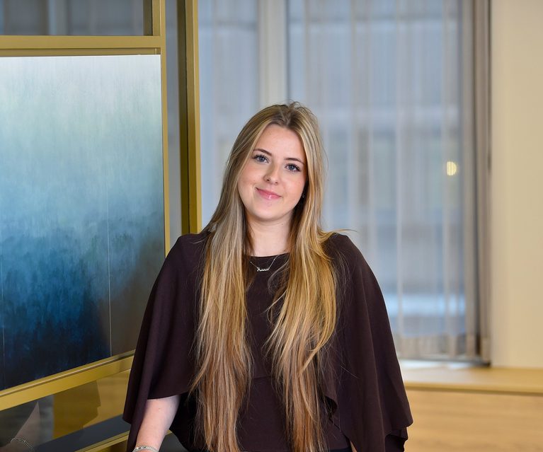 A young woman with long blonde hair, wearing a dark top, stands indoors near a window with blinds, smiling softly at the camera.