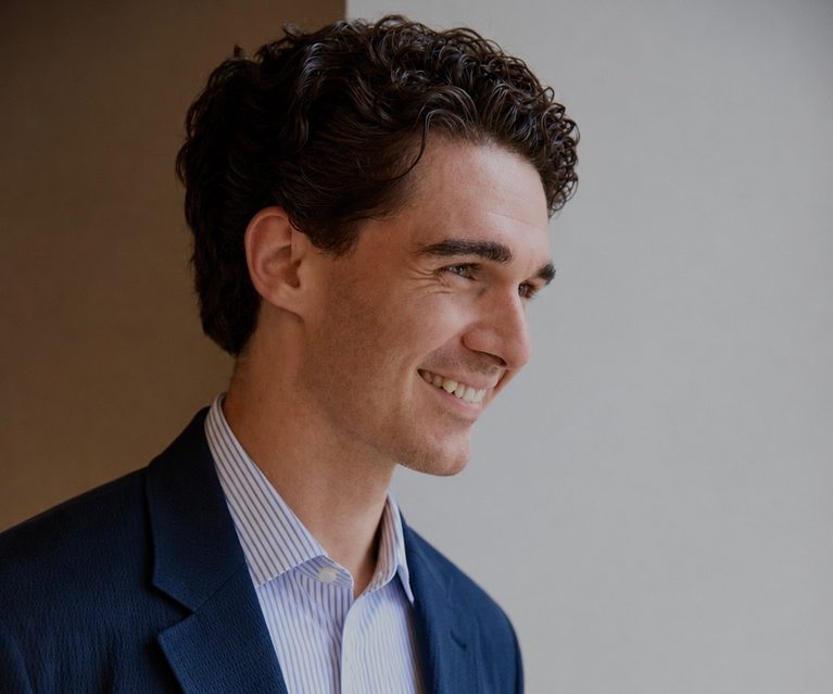 Side-profile of a person with dark, wavy hair in a navy blazer and pale striped shirt, smiling as they look to the right against a neutral wall.