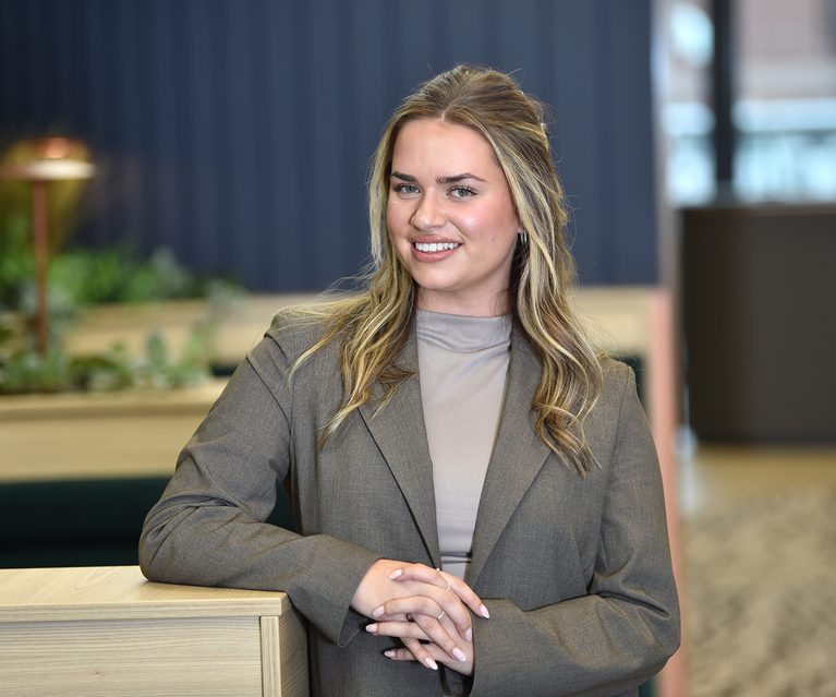 A young woman with blonde hair, wearing a grey blazer and light grey top, smiling and leaning on a wooden table in a modern, bright office setting.