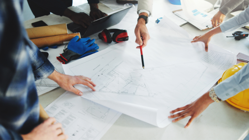 Team members review large architectural blueprints on a table, pointing with a pencil among laptops, gloves, a yellow hard hat, and tools.