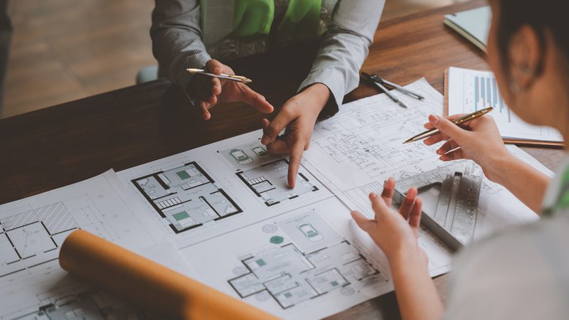 Two people discussing architectural plans at a table, with blueprints and drafting tools, in a workspace with a wooden floor.