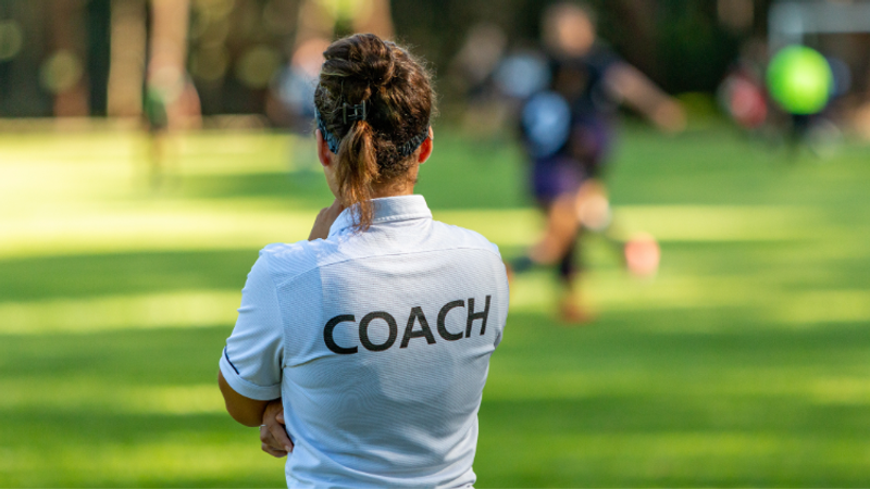 Person wearing a white polo labeled COACH stands with arms folded on a sunlit grassy sports field, facing away as blurred players practice and a game unfolds in the distance.