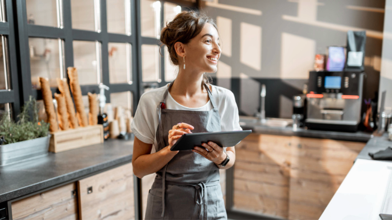 Person in a grey apron stands in a bright café kitchen, smiling while holding a tablet; loaves of bread on a wooden rack and a coffee machine in view behind.