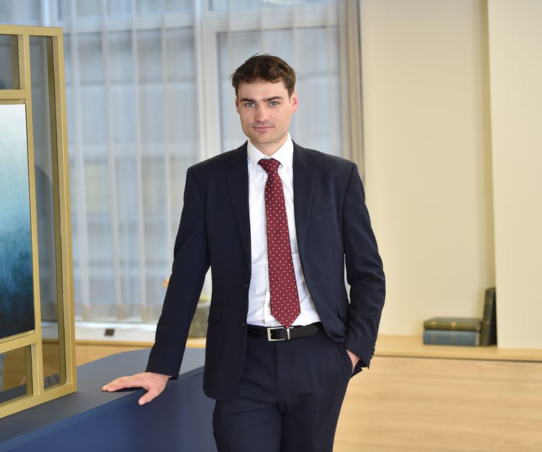 A man in a dark suit with a white shirt and red polka dot tie standing in an office, leaning on a desk with one hand in his pocket, in front of large windows with curtains.