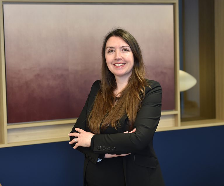 A woman with long brown hair wearing a black blazer smiles with crossed arms in an office setting.