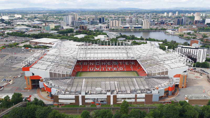 Aerial view of Old Trafford stadium, home of Manchester United, with red seating spelling MANCHESTER UNITED, and a white steel roof, set in a city by a river.