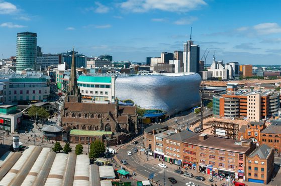 A cityscape with modern buildings, a historic church, a distinctive silver curved structure, and a busy street scene under a partly cloudy sky.