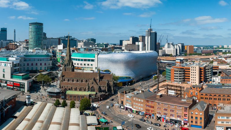 A cityscape with modern buildings, a historic church, a distinctive silver curved structure, and a busy street scene under a partly cloudy sky.