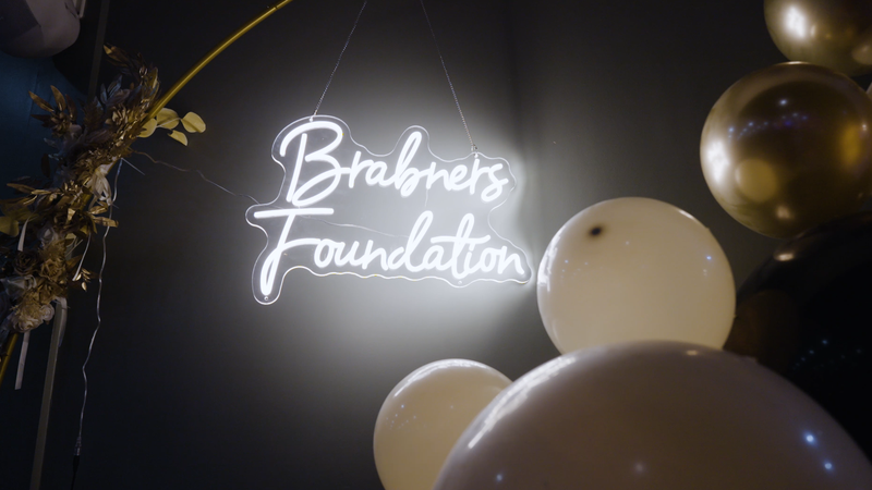A neon sign reads "Brabners Foundation" against a dark wall, surrounded by white and gold balloons, with some gold floral decorations to the left.