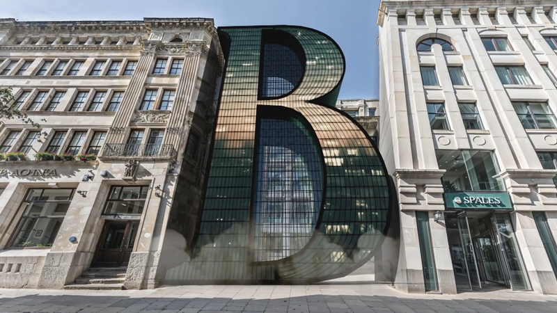 A large, modern glass sculpture of the letter 'B' is situated between two ornate, historic buildings on a city street, reflecting the surrounding architecture.