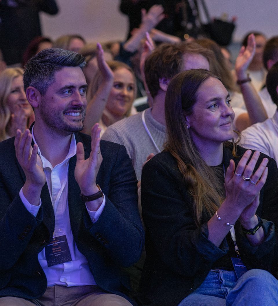 Audience at a conference sits in a dim room, clapping and smiling as cameras film in the background; attendees wear lanyards and a blue-lit backdrop surrounds them.