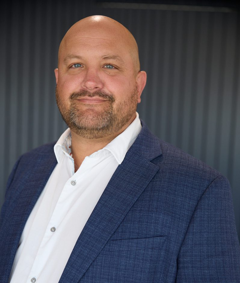Head-and-shoulder portrait of a bald person with blue eyes, wearing a white shirt and blue blazer, against a dark, vertically striped background.