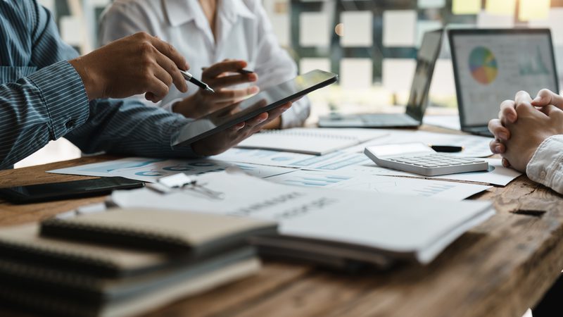 Two people sit at a wooden table with documents, a calculator, laptops displaying charts, and a tablet; they appear to be discussing business data.