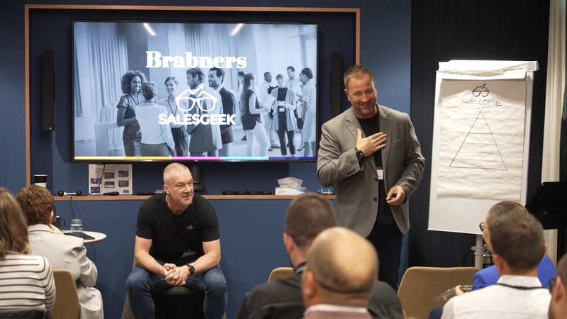 A man in a grey blazer is speaking and smiling to a group of people in a conference room. Some attendees are seated and facing him, with a large presentation screen and a flip chart behind him.