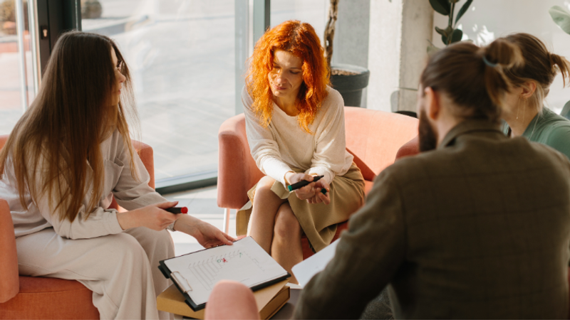 Four people sit on pink chairs and a sofa in a bright room with large windows; they are engaged in a discussion, with notebooks and papers on a small table.
