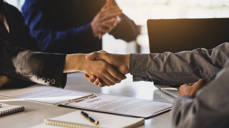Two people shaking hands across a desk with documents and writing materials, while a third person in the background appears to be clapping or gesturing.