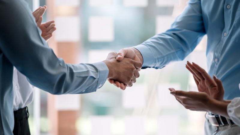 Two people are shaking hands in a professional setting, with a woman clapping nearby and blurred windows in the background.