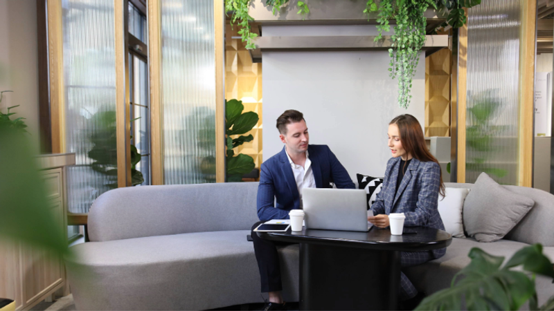 A man and woman sit together on a grey sectional sofa in a modern, well-lit room with greenery, looking at a tablet and working on a laptop on a small black coffee table.