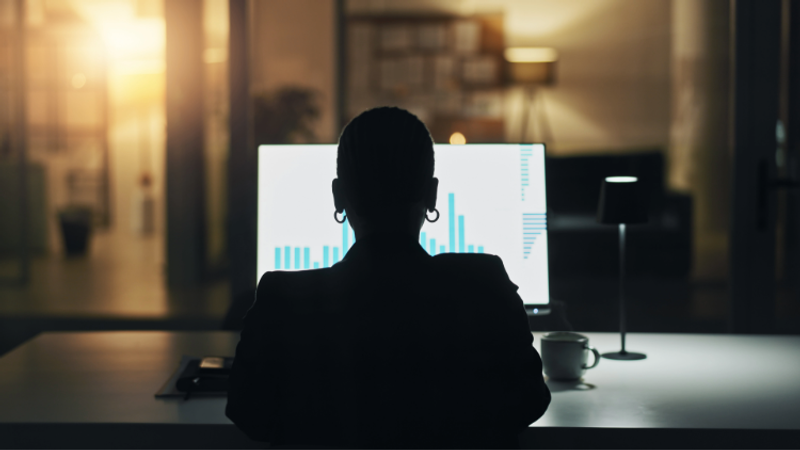 A person with short hair and hoop earrings sitting at a desk in front of a computer displaying bar graphs, in a dimly lit room with warm background lighting.