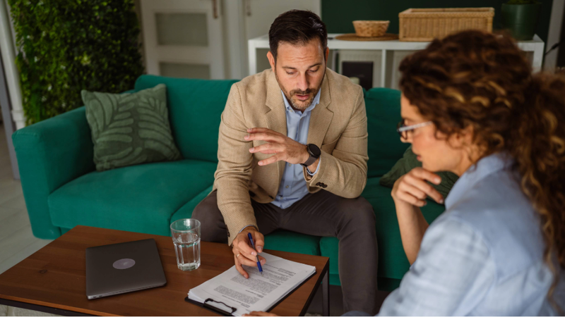 A man and woman having a discussion in a living room, with documents, a laptop, and a glass of water on the table between them.