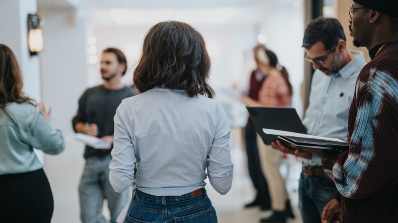 A group of diverse people engaging in conversation at an indoor professional networking event or seminar, with some participants holding notebooks or tablets.