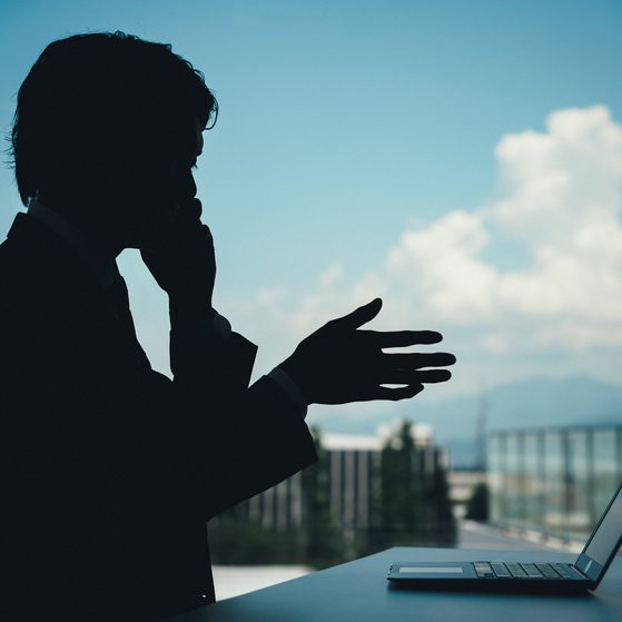 Silhouette of a person in front of a window with a laptop on a table, with a cityscape and sky with clouds in the background.