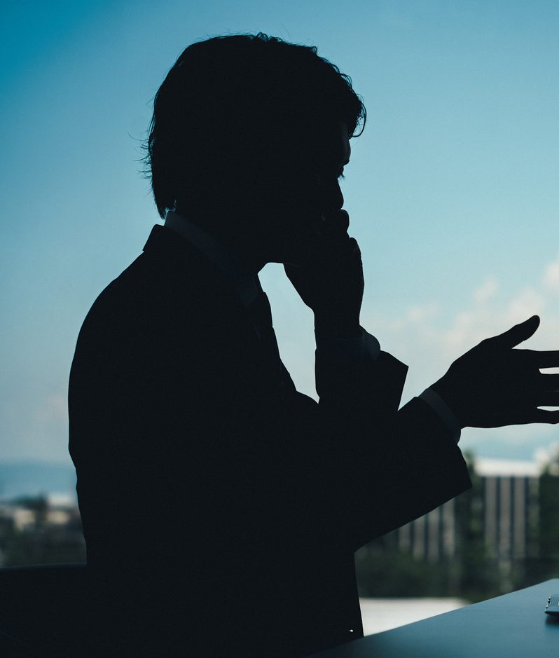 Silhouette of a person in front of a window with a laptop on a table, with a cityscape and sky with clouds in the background.