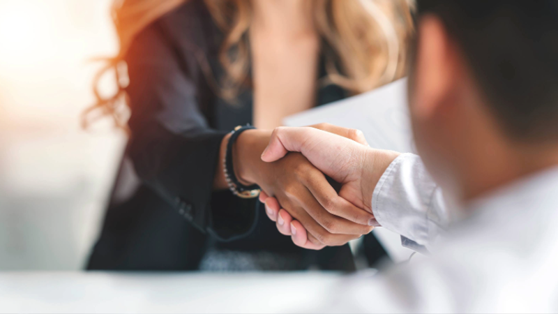 Close-up of two people shaking hands, with a woman holding some papers in the background, in a professional setting.