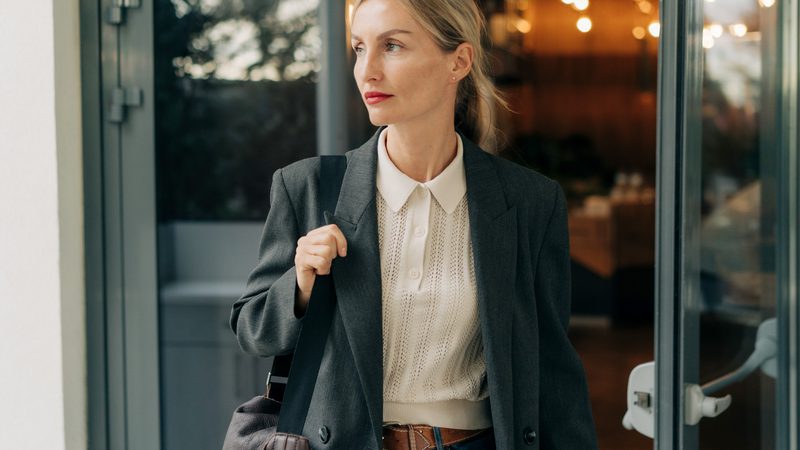A woman in a dark grey blazer, white knit top, and jeans holding a black shoulder bag, standing near a glass door with a thoughtful expression.