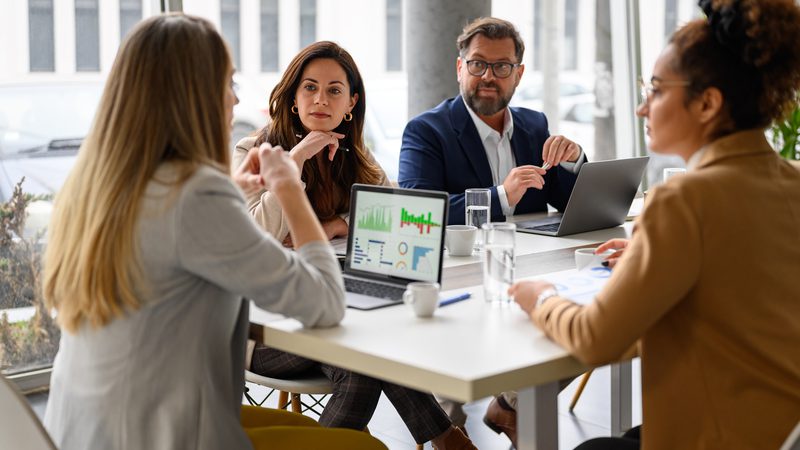 Four professionals are seated around a table in a modern office, engaged in a discussion, with laptops and glasses of water in front of them.