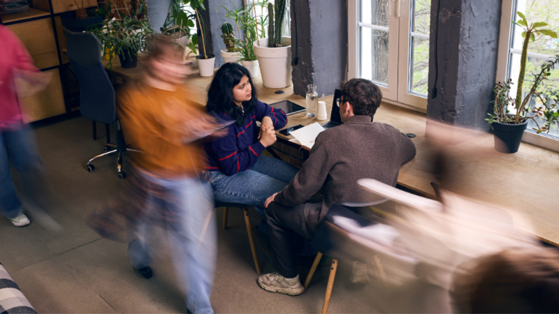 Two people sit at a wooden table by large windows, mid-conversation, while several others blur past in a busy, plant-filled indoor space bathed in natural light.