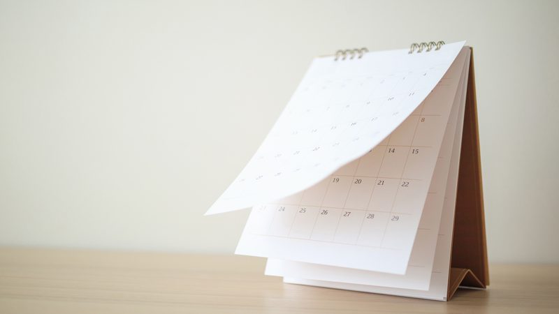 A slim white desk calendar on a light wooden surface, held open by a metal spiral binding, showing a grid of dates in a minimal monthly layout.