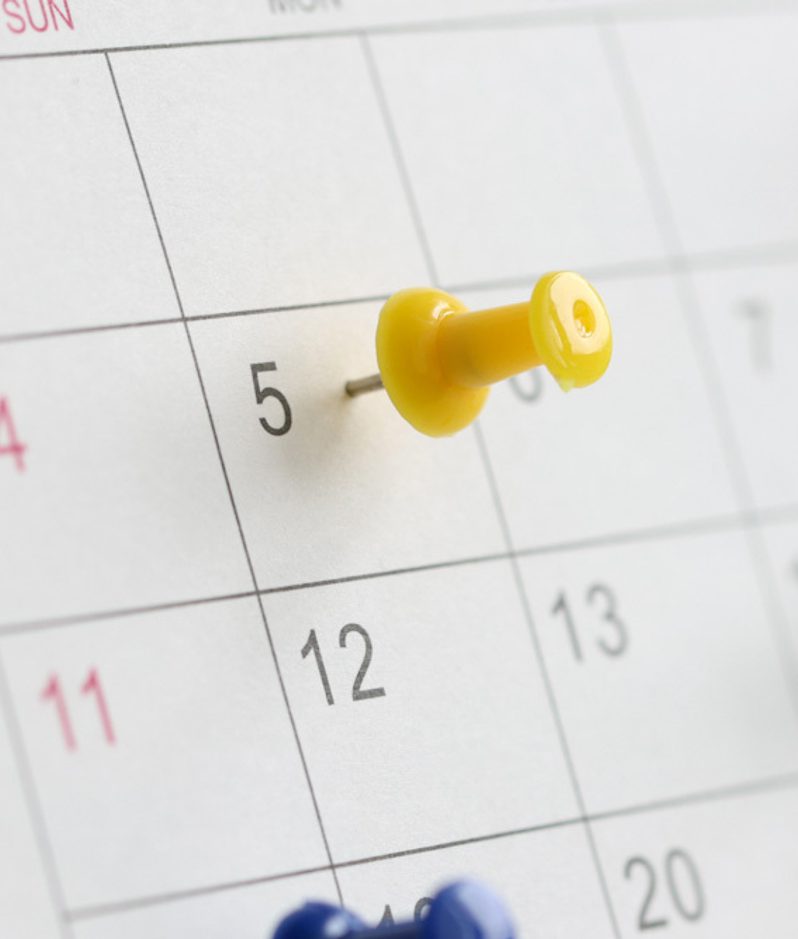 Close-up of a calendar page with grid lines and multiple coloured push pins marking dates; a yellow pin on the 5th, others blurred in the background.