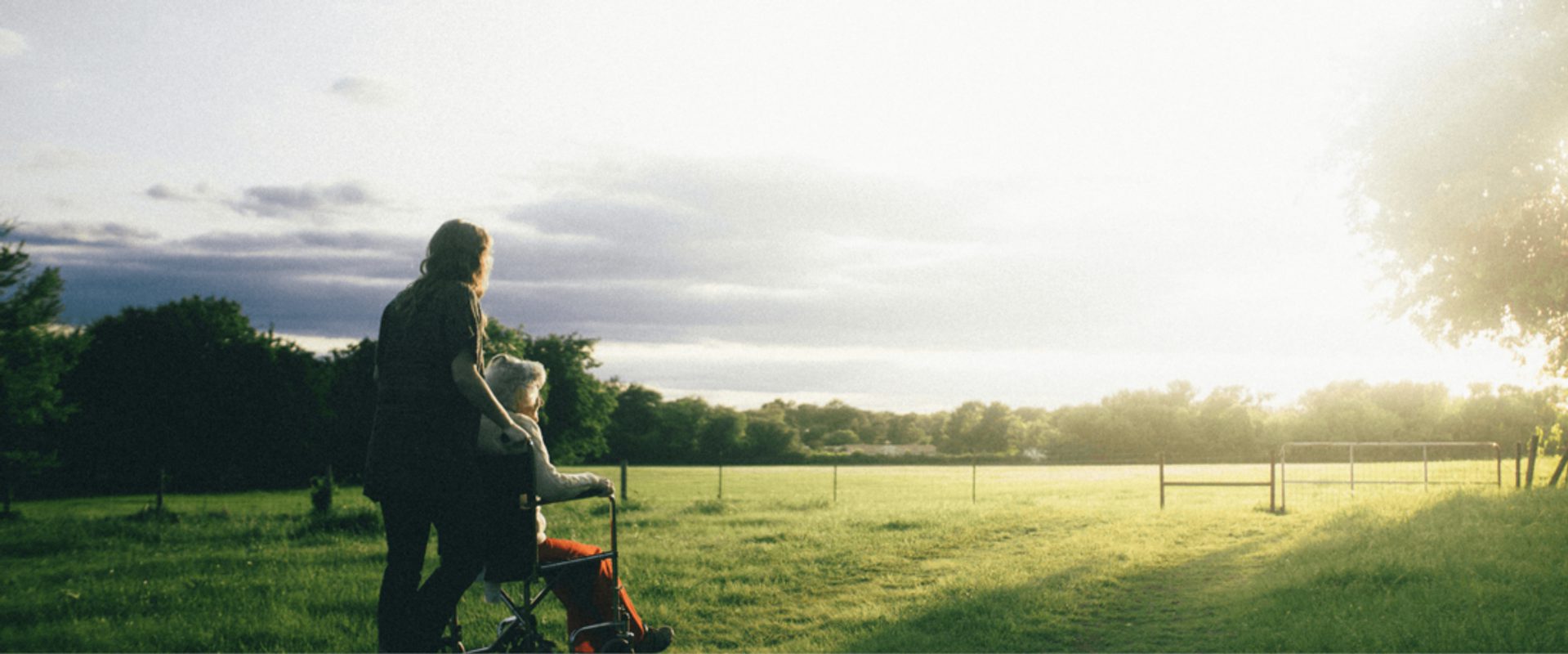 A woman with a child in a stroller walking across an open grassy field during sunset, with trees and fencing visible in the background.